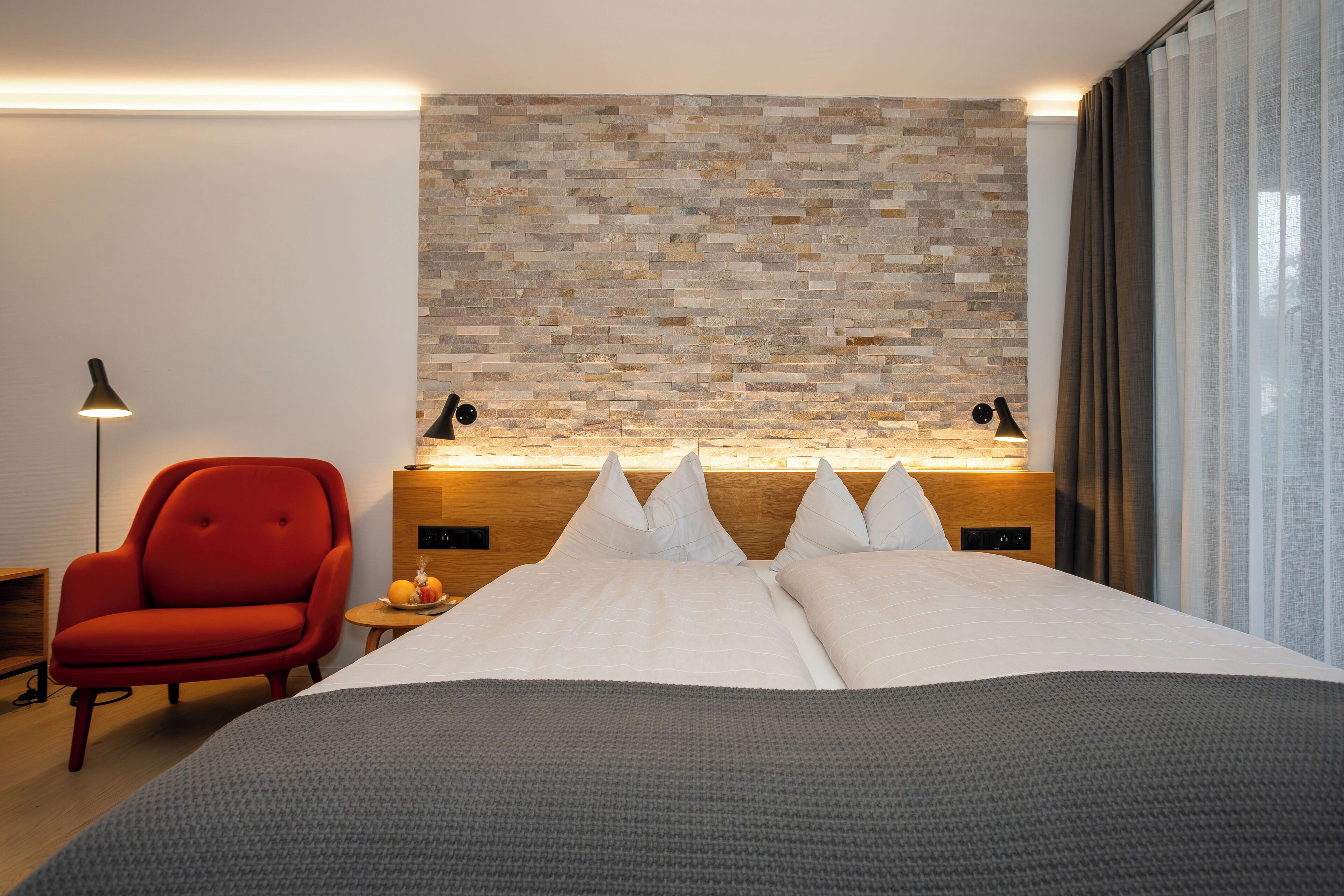 A modern hotel room with a large bed, a red armchair, a small table with a lamp, and a stone-patterned wall.