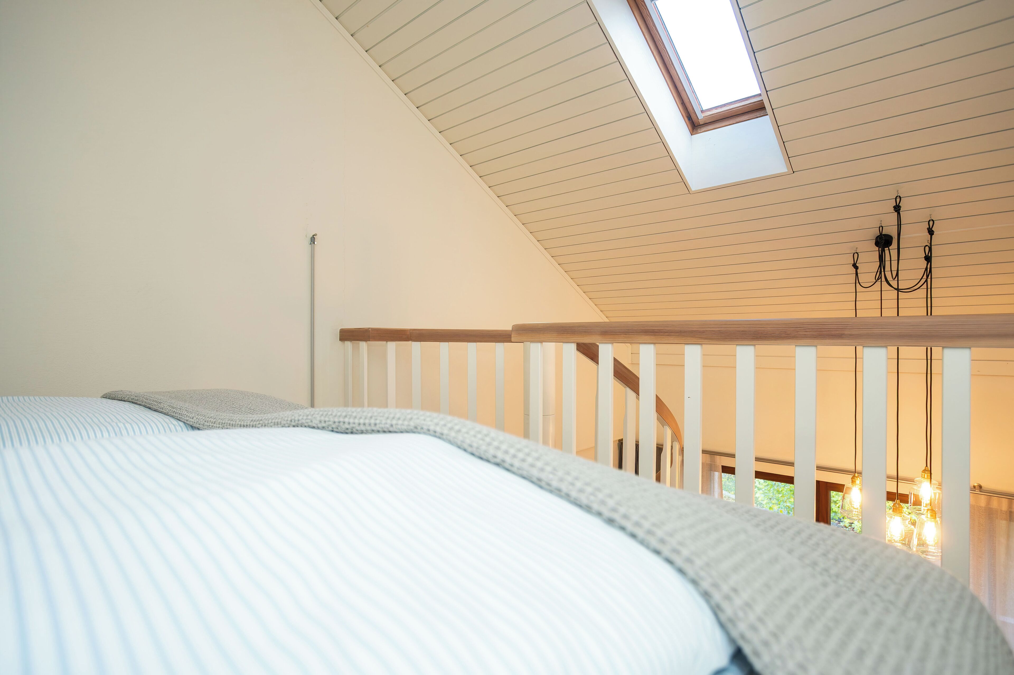 Ein Schlafzimmer mit schrägem Dach, einem Oberlicht, einem Bett mit gestreifter Decke, einer Holzgalerie und hängenden Lampen.