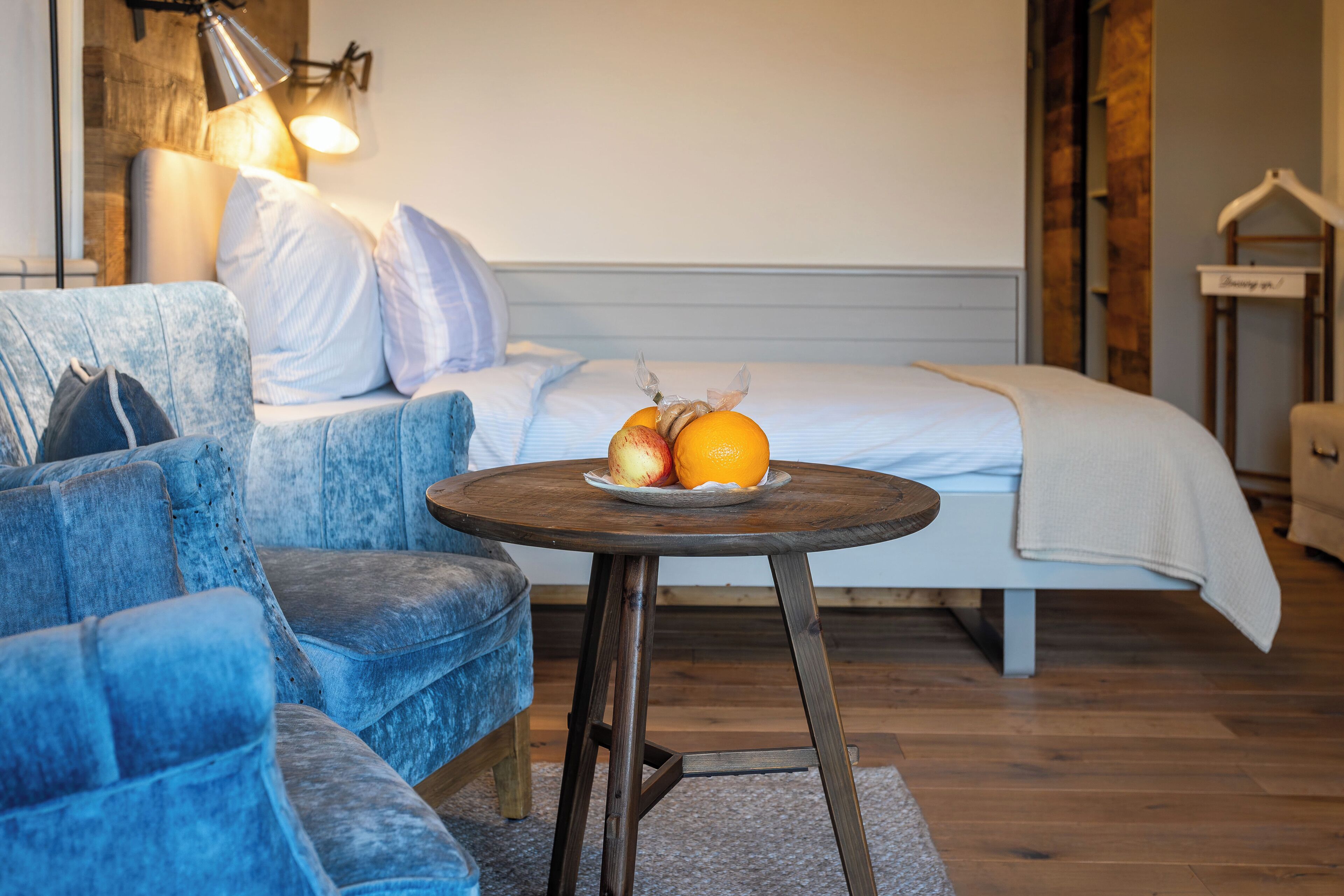 Ein gemütliches Zimmer mit einem blauen Sofa, einem kleinen runden Tisch mit Obst, einem Bett mit weißer Bettwäsche und einer hängenden Lampenleuchte.