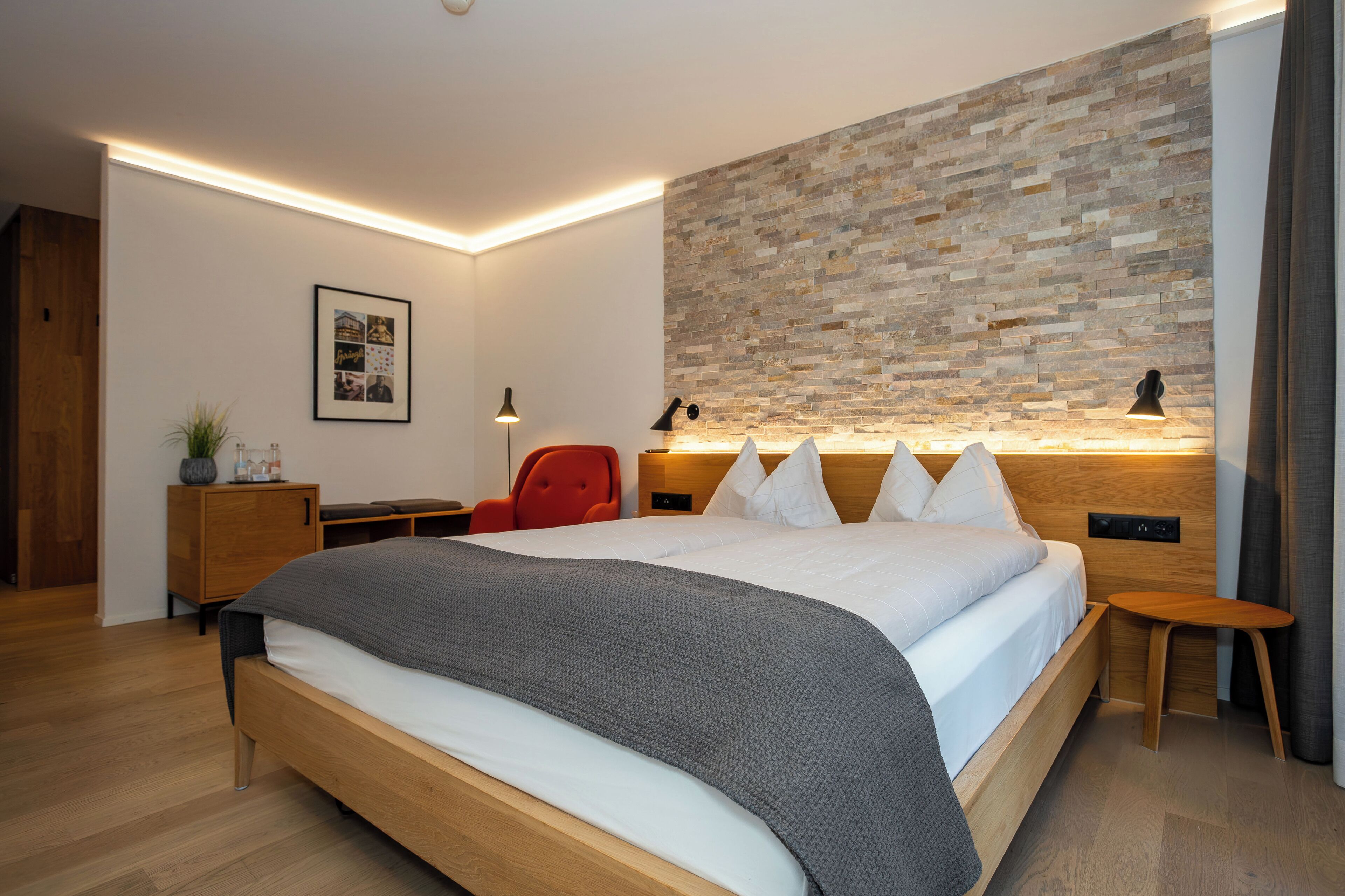 Ein modernes Schlafzimmer mit einem großen Bett, einem Holz-Kopfteil, einem roten Sessel, einem Nachttisch und einer Wand mit einer strukturierten Oberfläche.