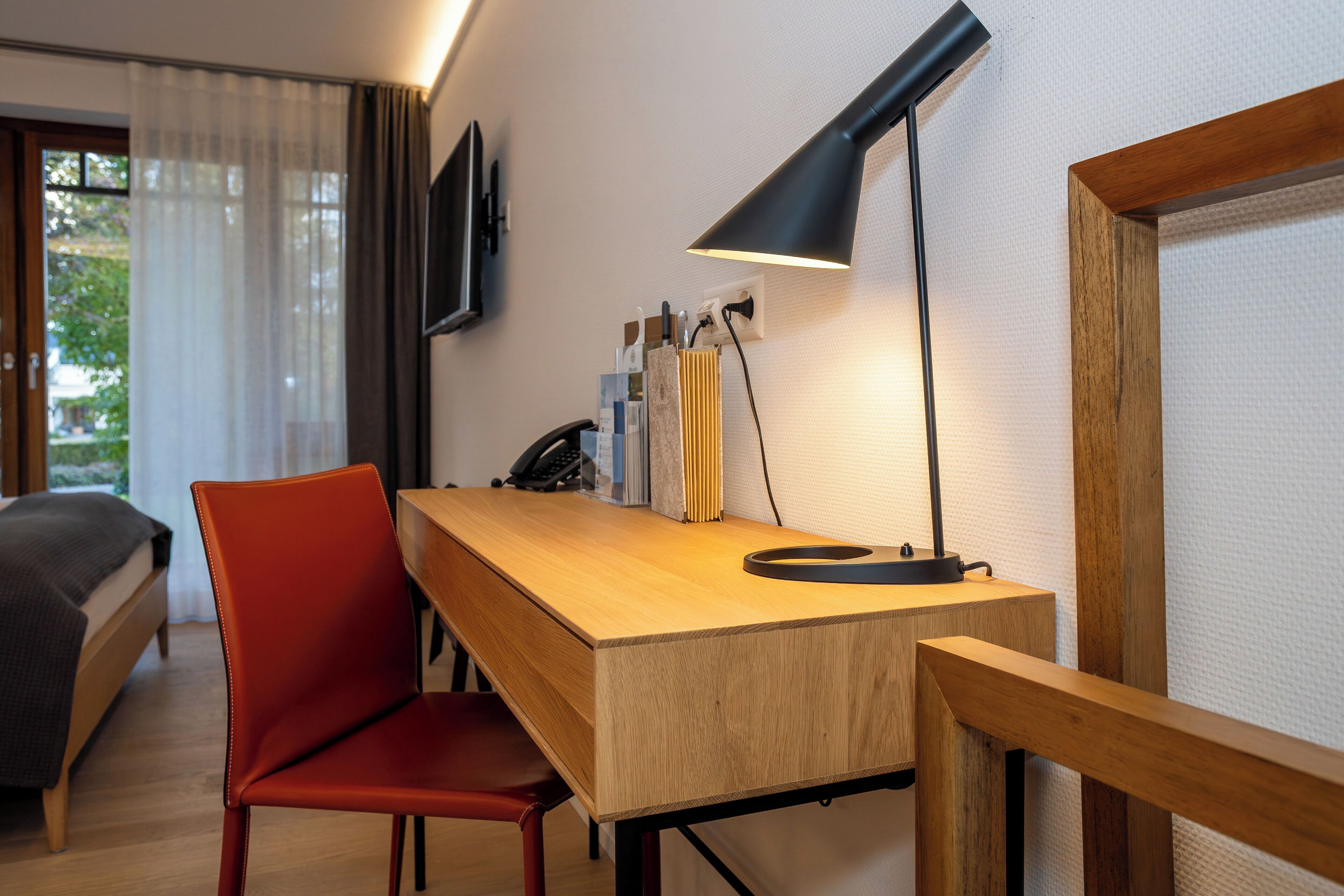 A hotel room with a wooden desk, a red chair, a bed, a TV mounted on the wall, and a lamp.