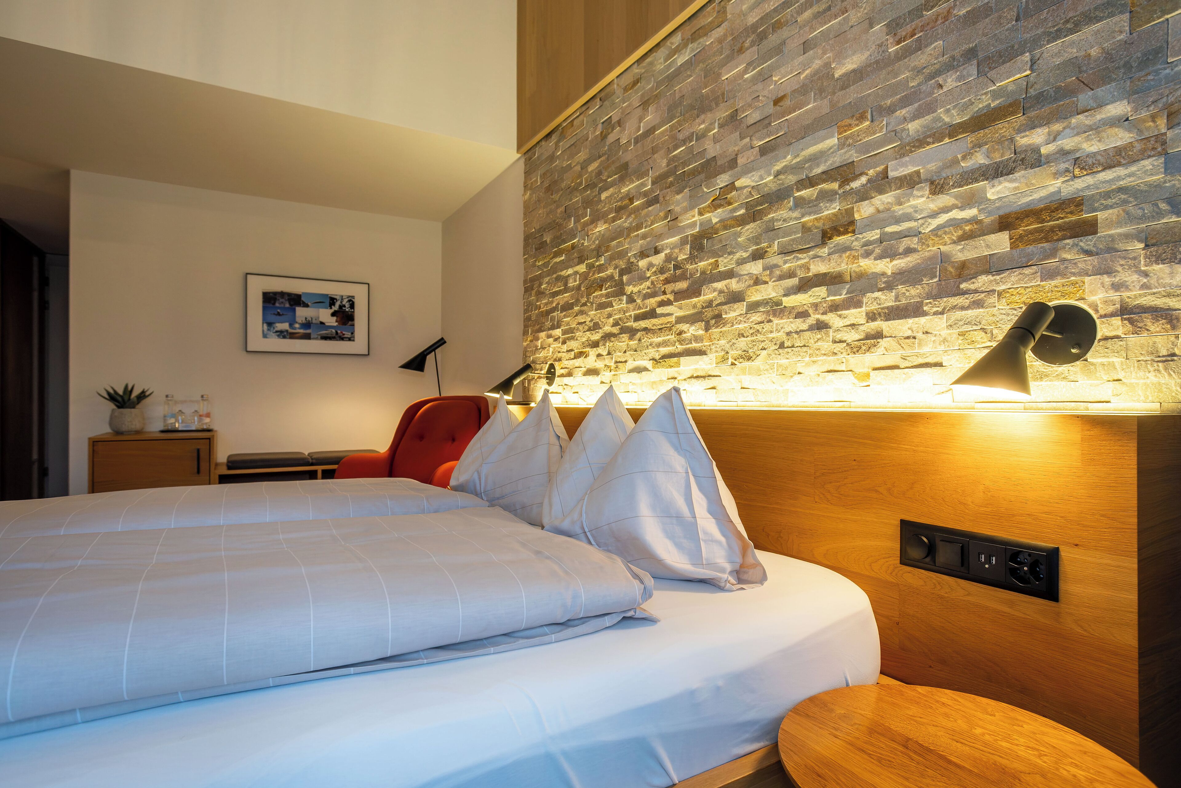 A modern hotel room with a large bed, a wooden headboard, a stone wall, a bedside table with a lamp, and a small desk with a chair.
