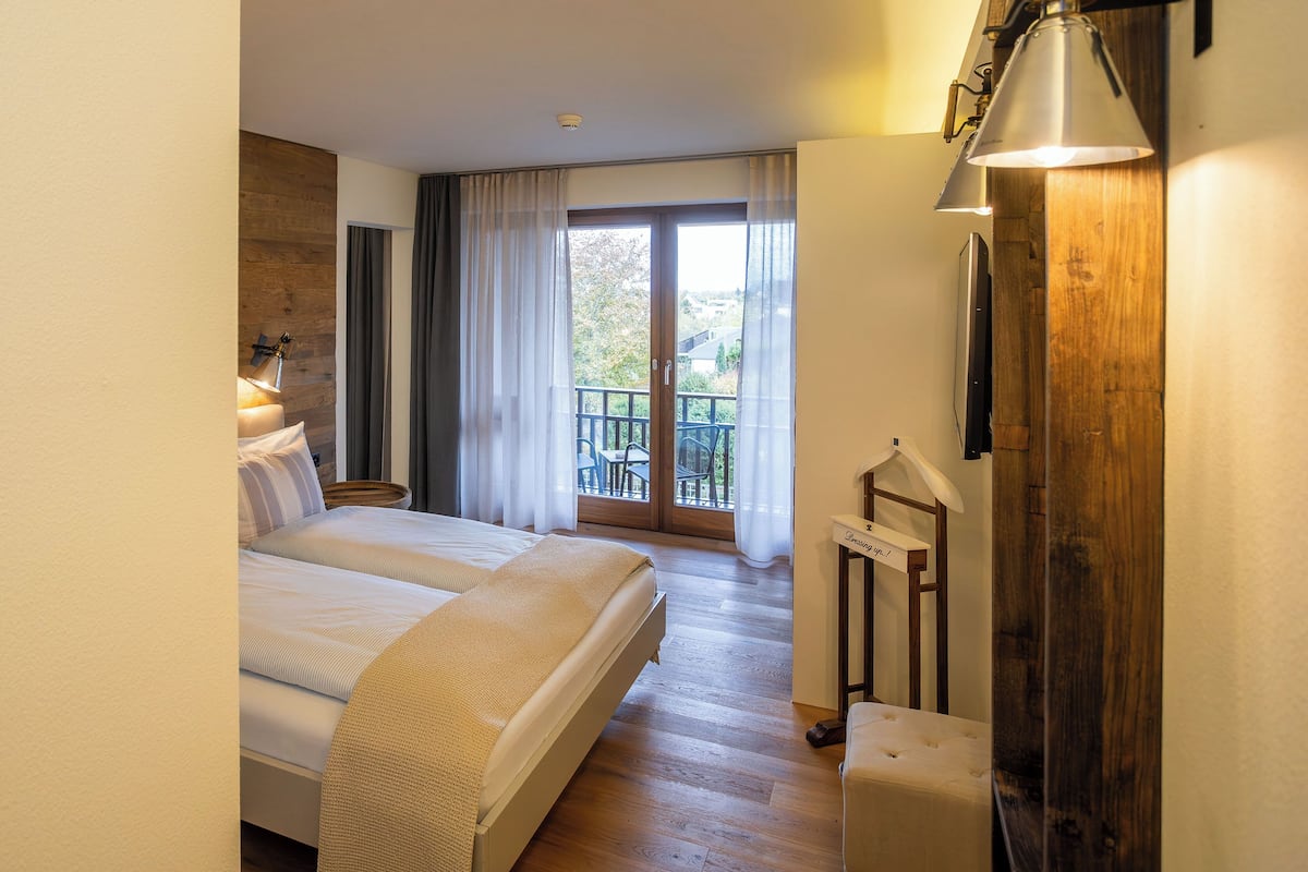 A hotel room with a large bed, a wooden floor, a balcony with a view, and a mounted TV.