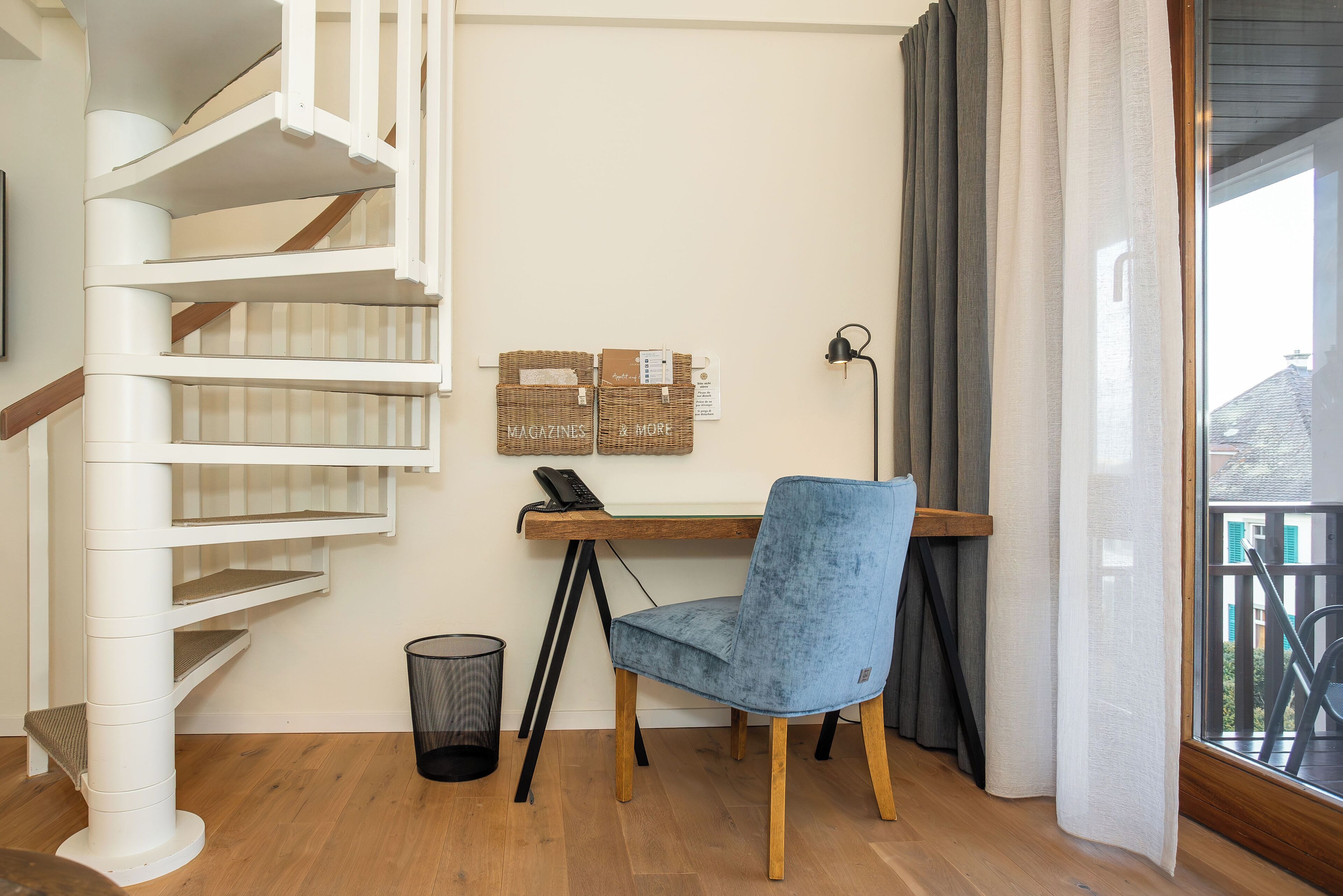 A modern room with a spiral staircase, a desk with a chair, a trash bin, and a window with curtains.