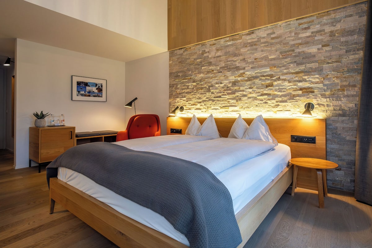 A modern bedroom with a wooden bed, a red chair, a small wooden table, a nightstand, and a wall-mounted lamp.