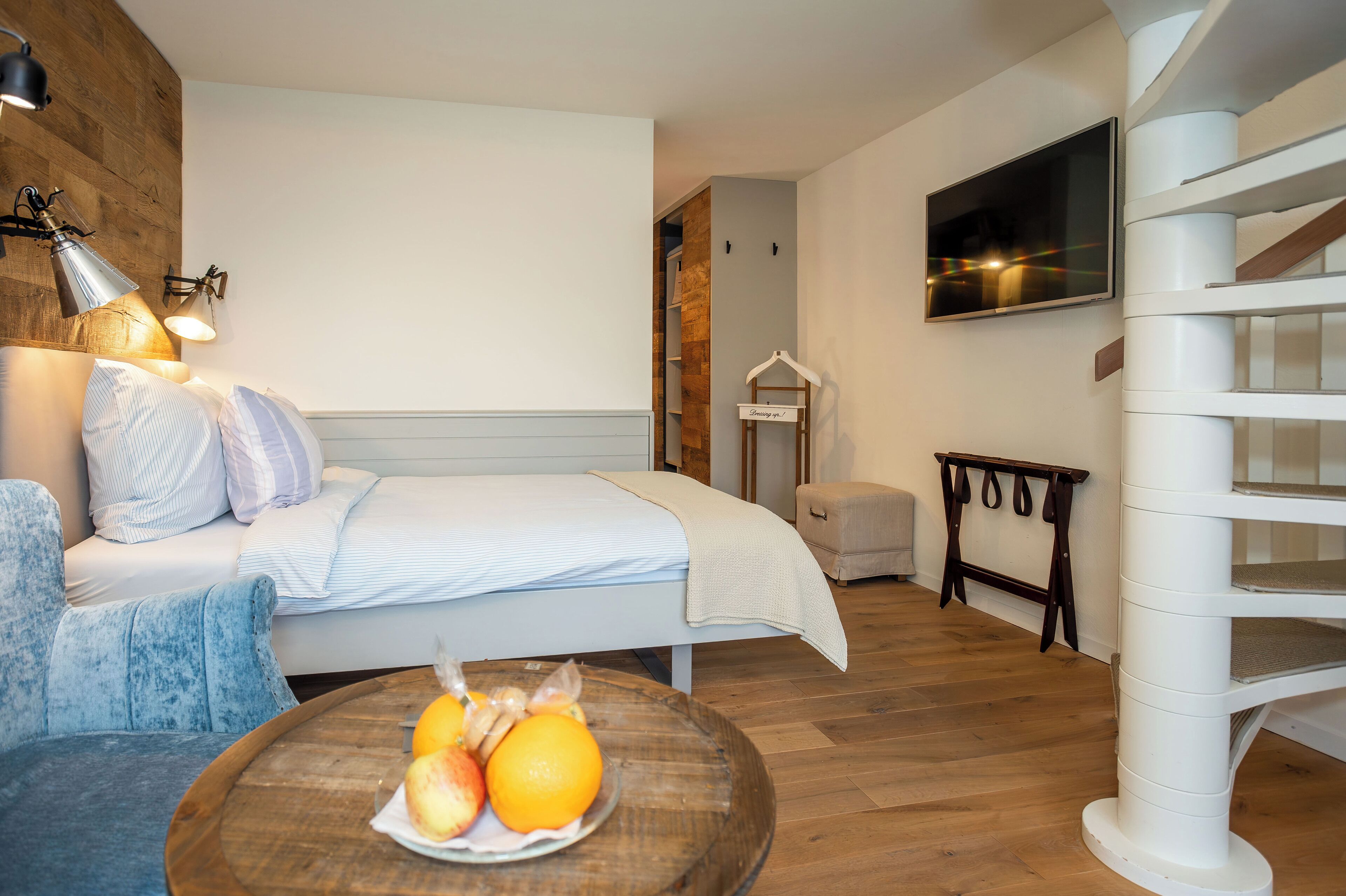 A bedroom with a bed, bedside table with fruit, a hanging coat rack, and a staircase.