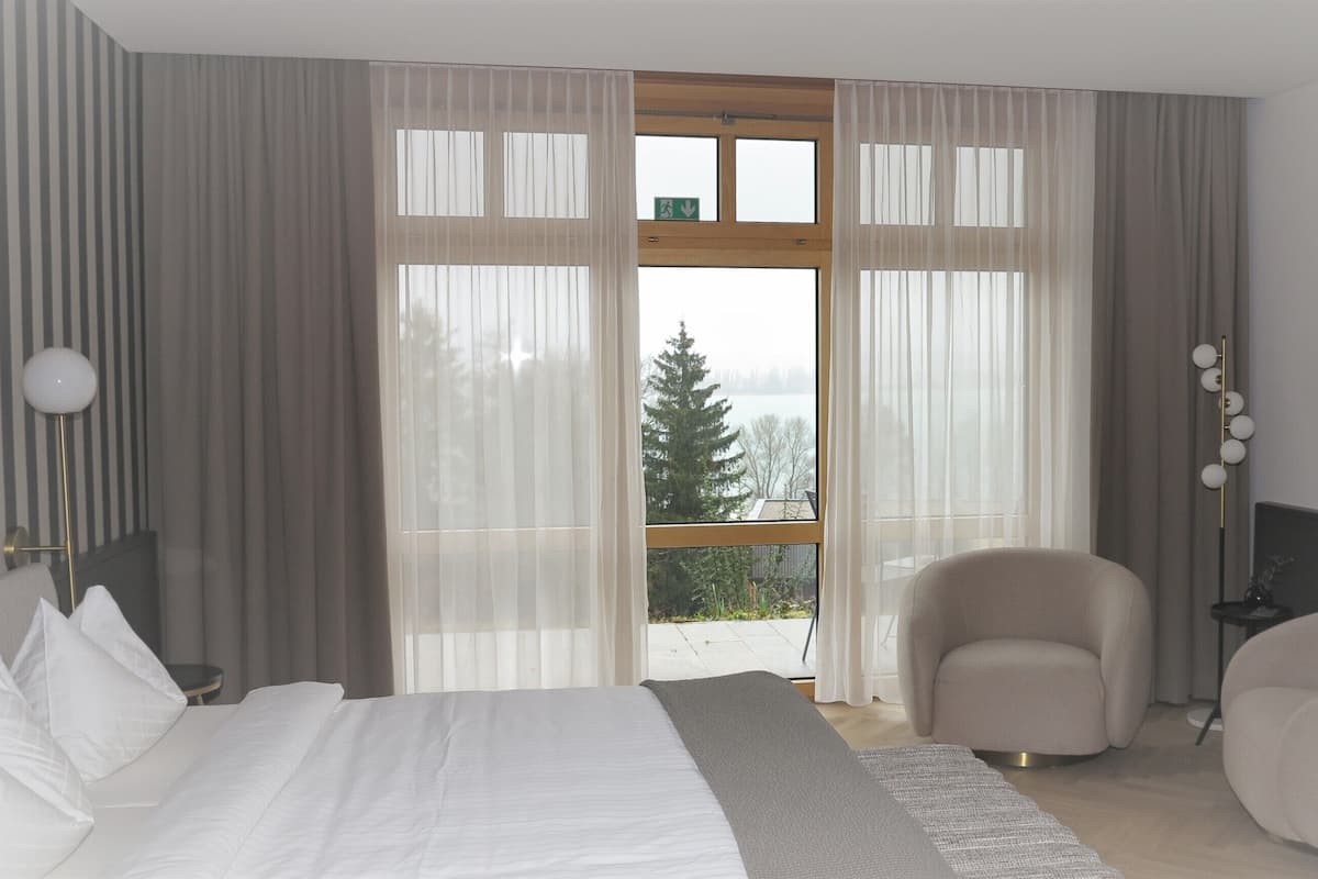 A modern bedroom with a large bed, a chair, and a view of a forest through a large window.