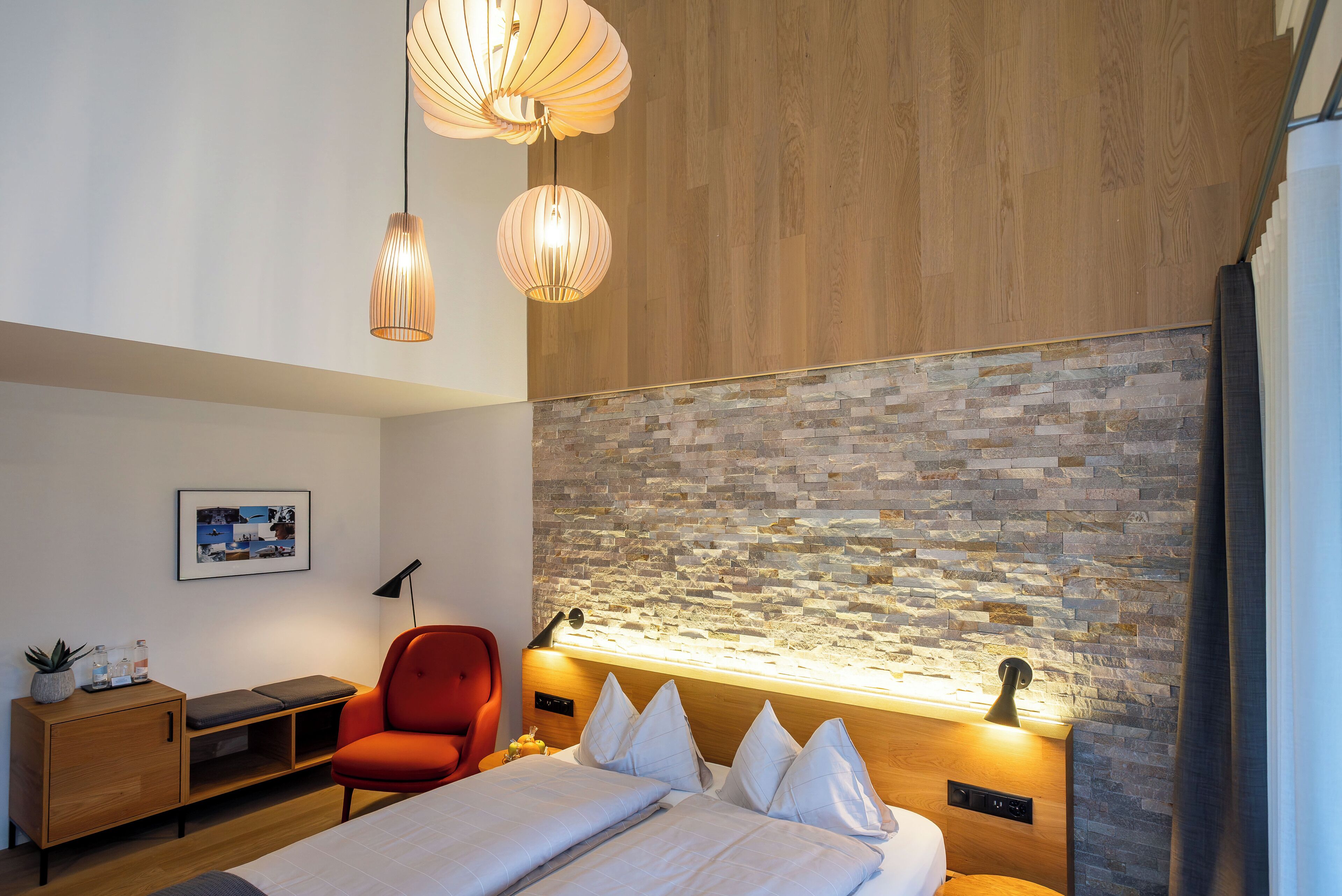 A modern hotel room with a wooden ceiling, stone wall, and a bed with white linens. There is a red chair and a small wooden cabinet with a plant.