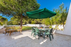 Un patio al aire libre con una mesa y sillas, un parasol verde y vistas a árboles y una pared blanca.