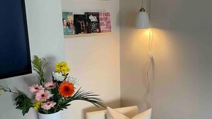 A modern living room with a white armchair, a small table with a vase of flowers, a wall-mounted TV, and a shelf with books and decorative items.