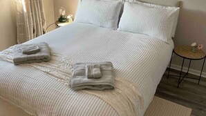 A neatly made bed with pillows and folded towels, a bedside table with a lamp, and a small round table with a flower vase.