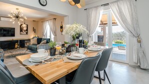 A modern dining area with a wooden table set for four, surrounded by grey chairs. A large window with curtains allows natural light to fill the room. - Villa Paula (Privlaka)