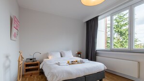 Un dormitorio ordenado con cama, mesita de noche y ventana con vistas a árboles.