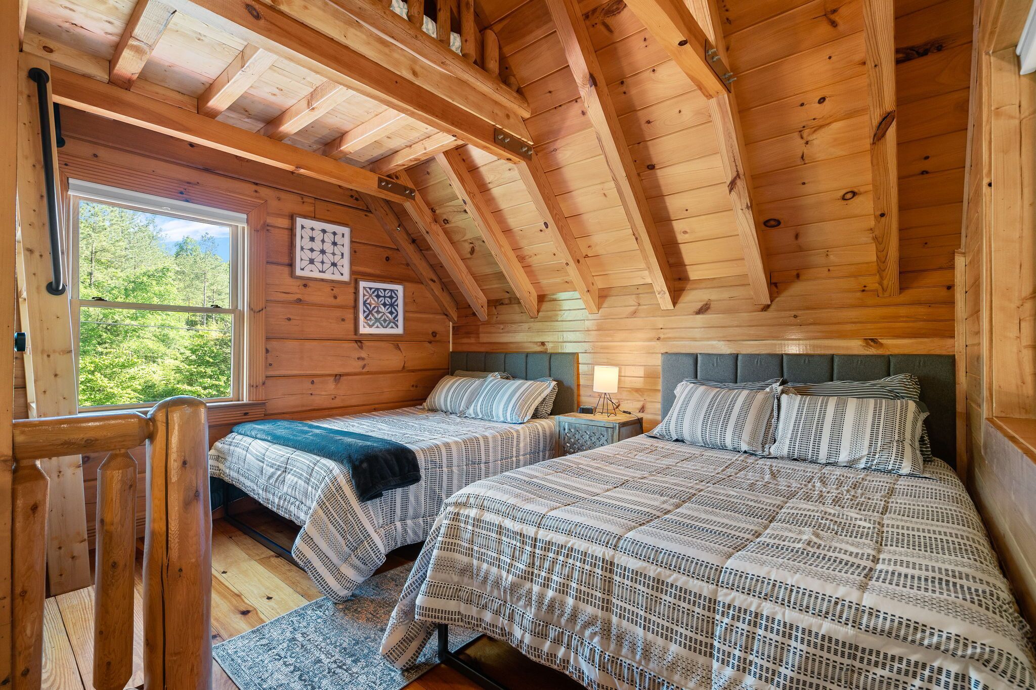 A cabin with two beds, wooden walls, a sloped ceiling, and a window with a view of trees.