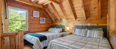 A cabin with two beds, wooden walls, a sloped ceiling, and a window with a view of trees.