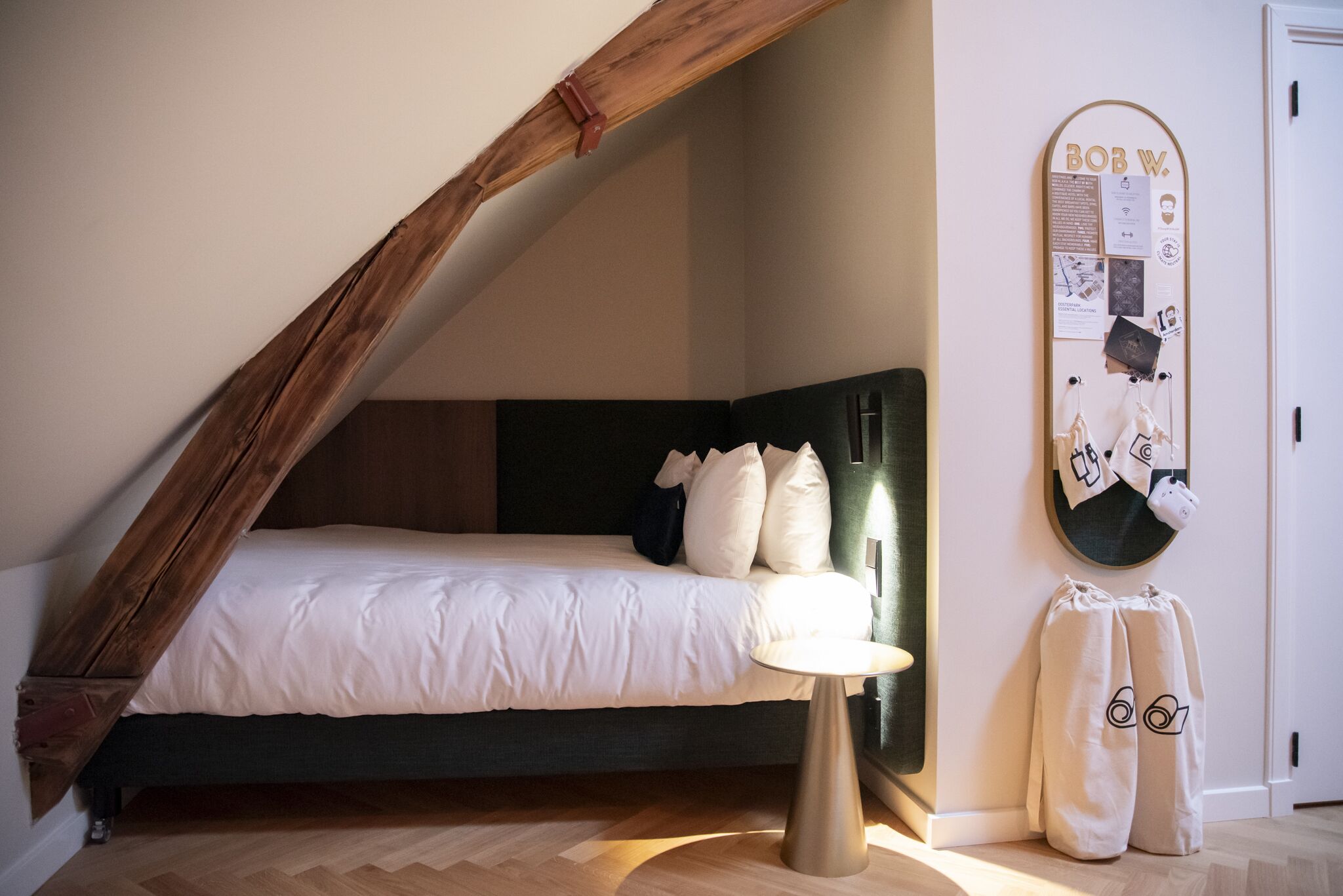 A bedroom with a sloped ceiling, a bed with white bedding, a bedside table, and a bulletin board with various items pinned to it.