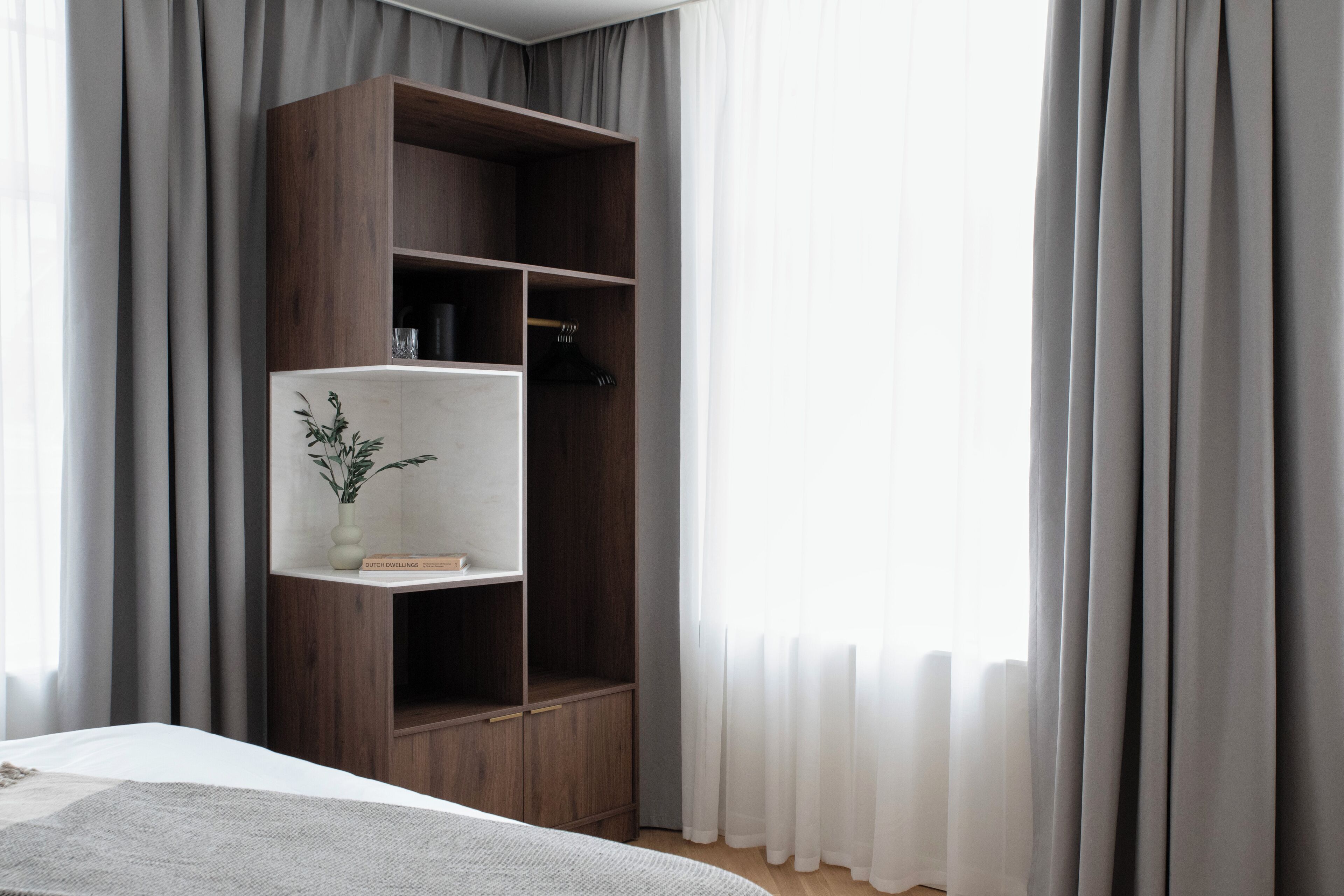 Une chambre à coucher avec un lit, une armoire en bois, une petite étagère avec une plante et des rideaux légers.