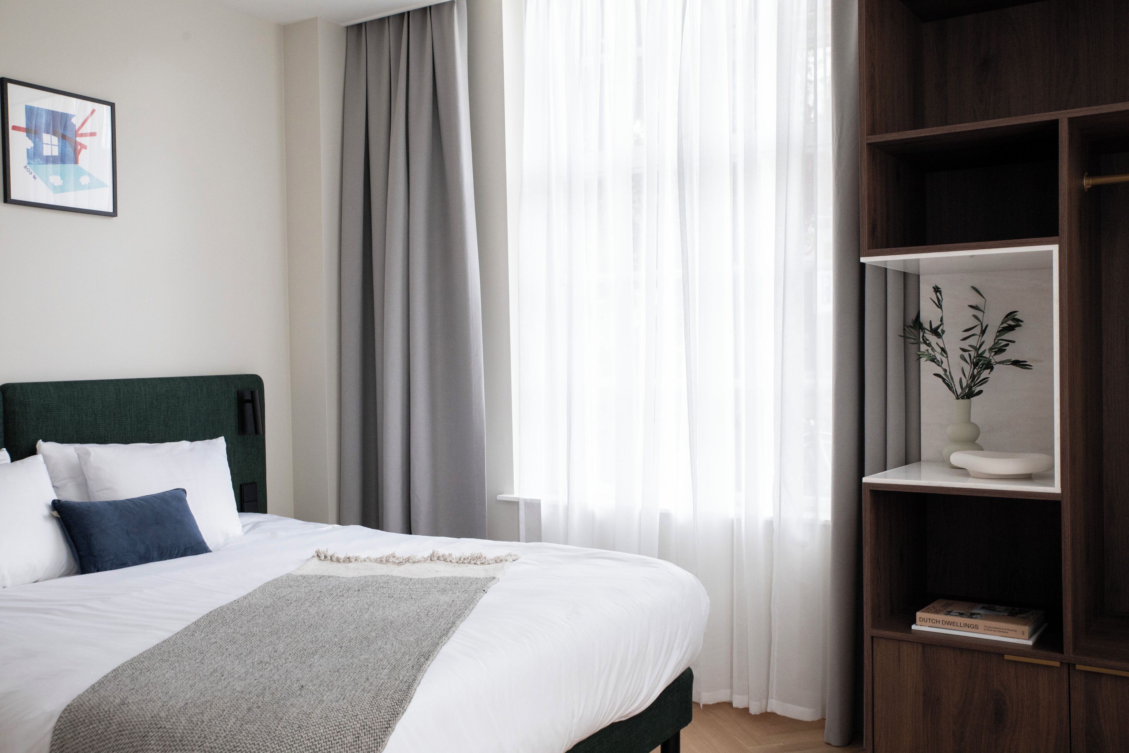 Ein ordentlich bezogenes Bett mit weißer und blauer Bettwäsche, einer grauen Decke und einem hölzernen Kopfteil. Rechts ist ein Bücherregal mit einer Pflanze und einem Buch zu sehen.