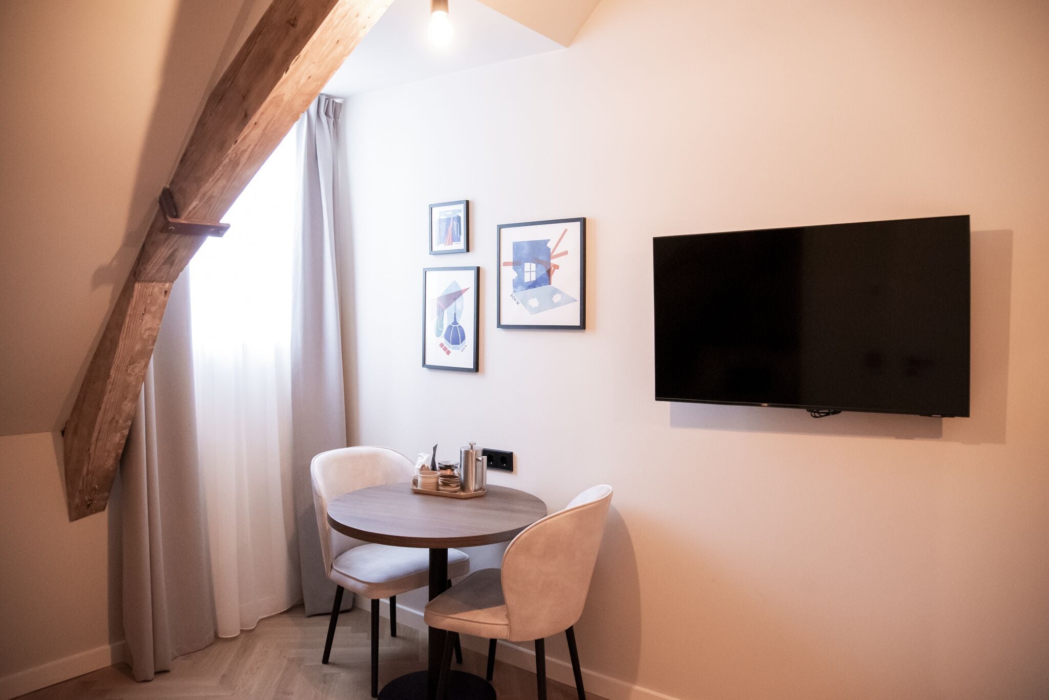 Een kleine, moderne kamer met een ronde tafel en twee stoelen, een flatscreen televisie aan de muur en een raam met gordijnen.
