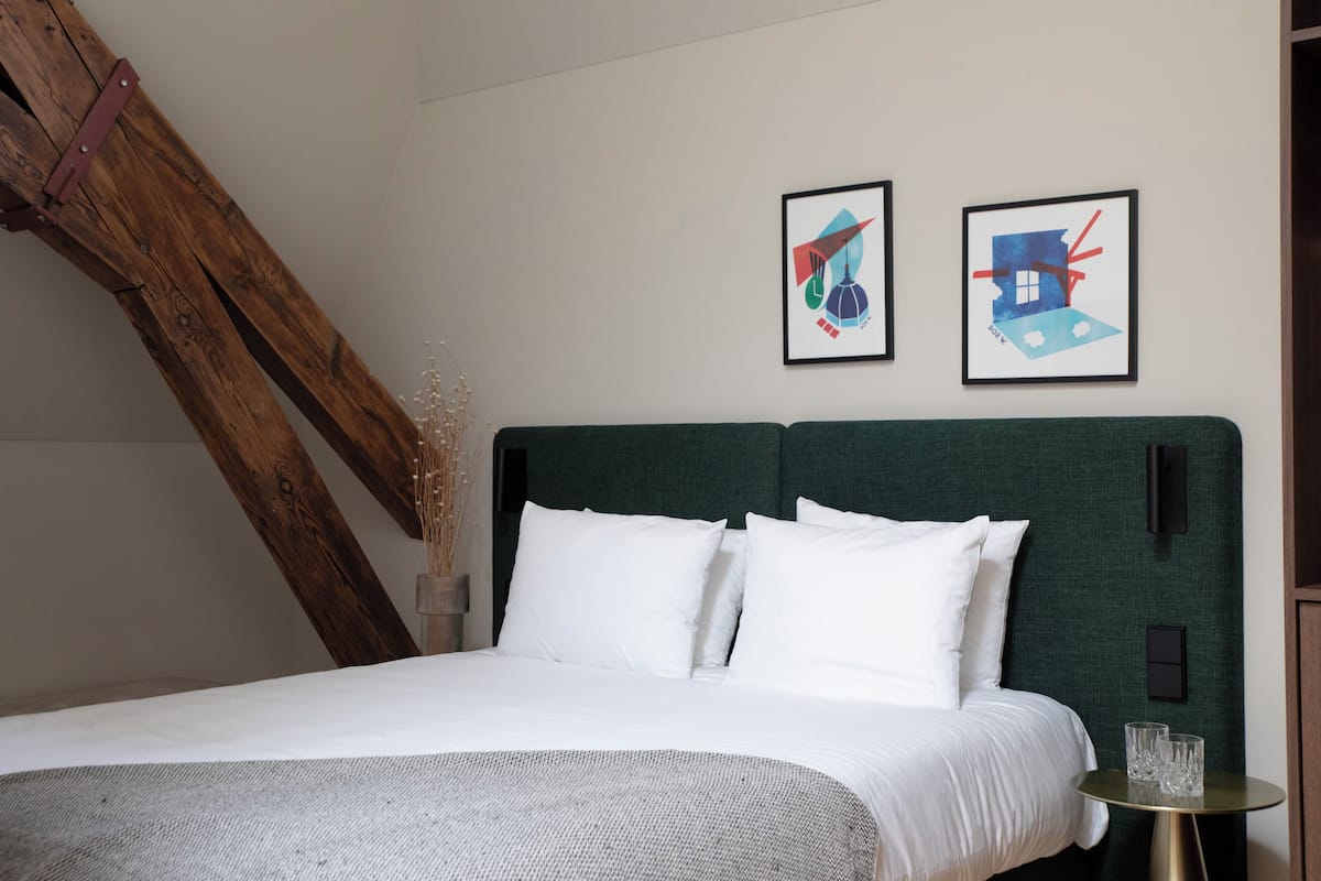 A neatly made bed with white linens and a green headboard, two framed artworks on the wall, and a small round table with glasses.