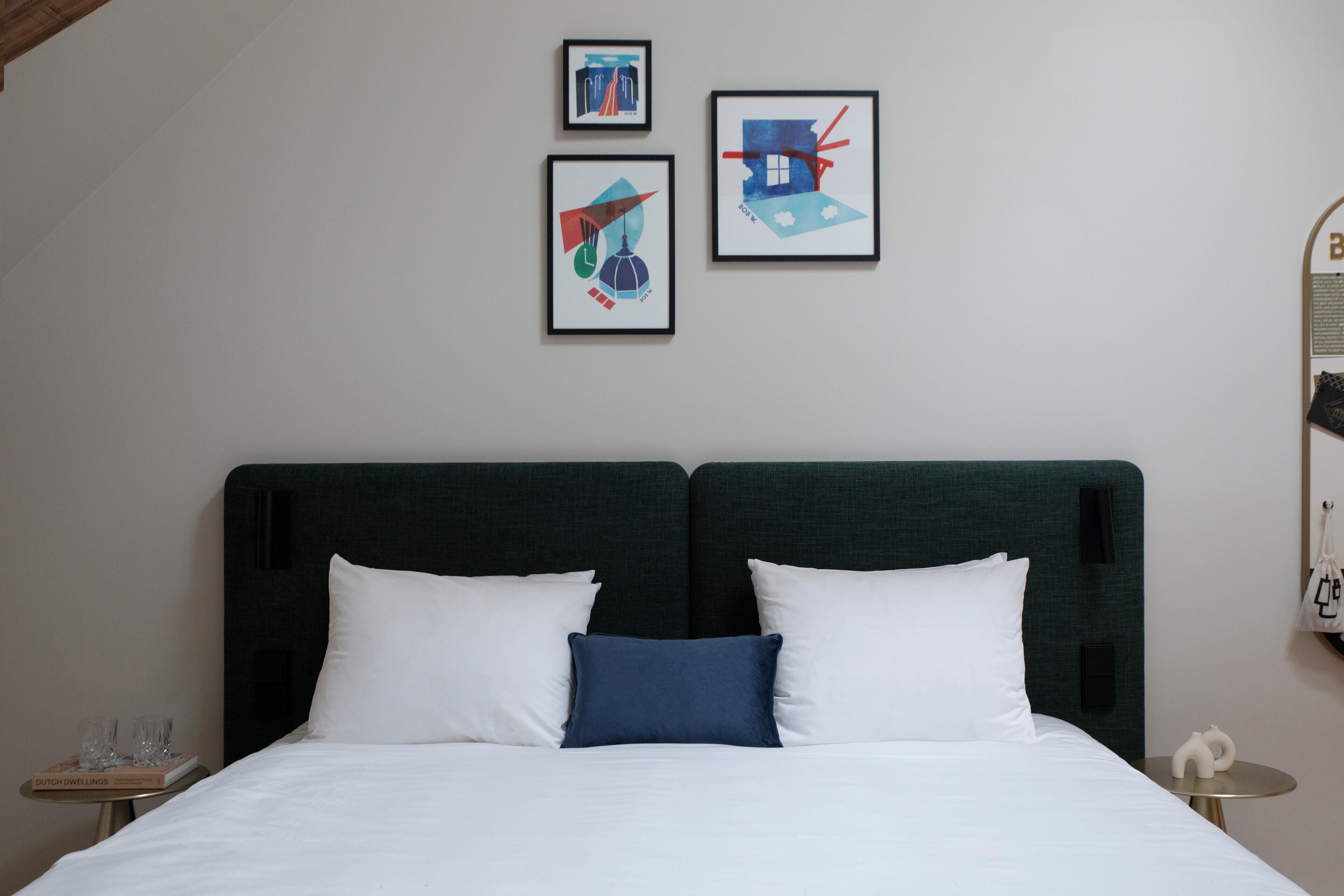 A neatly made bed with white and blue pillows, a dark green headboard, and three framed artworks on the wall.