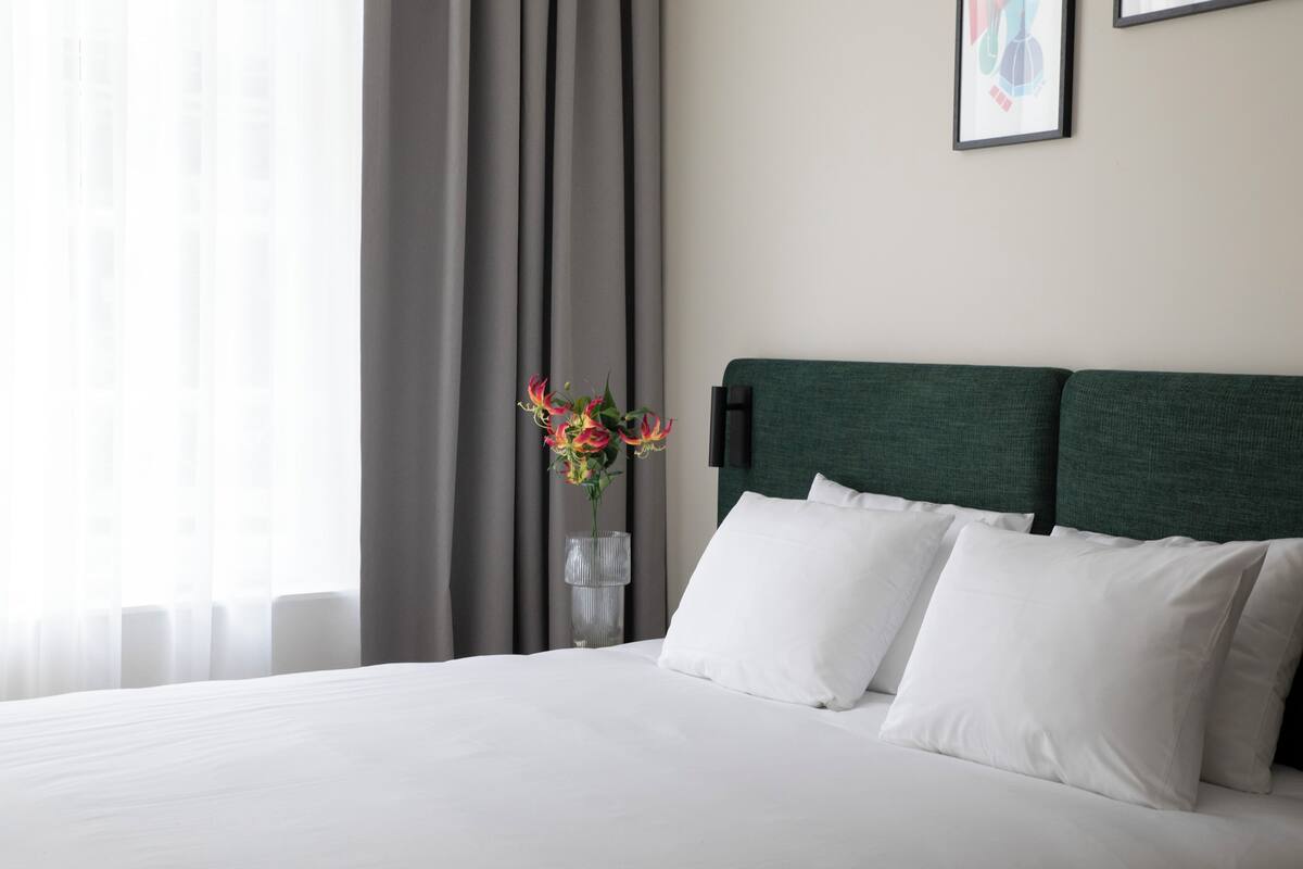 A hotel room with a bed, white pillows, a green headboard, a vase with flowers, and a framed picture on the wall.