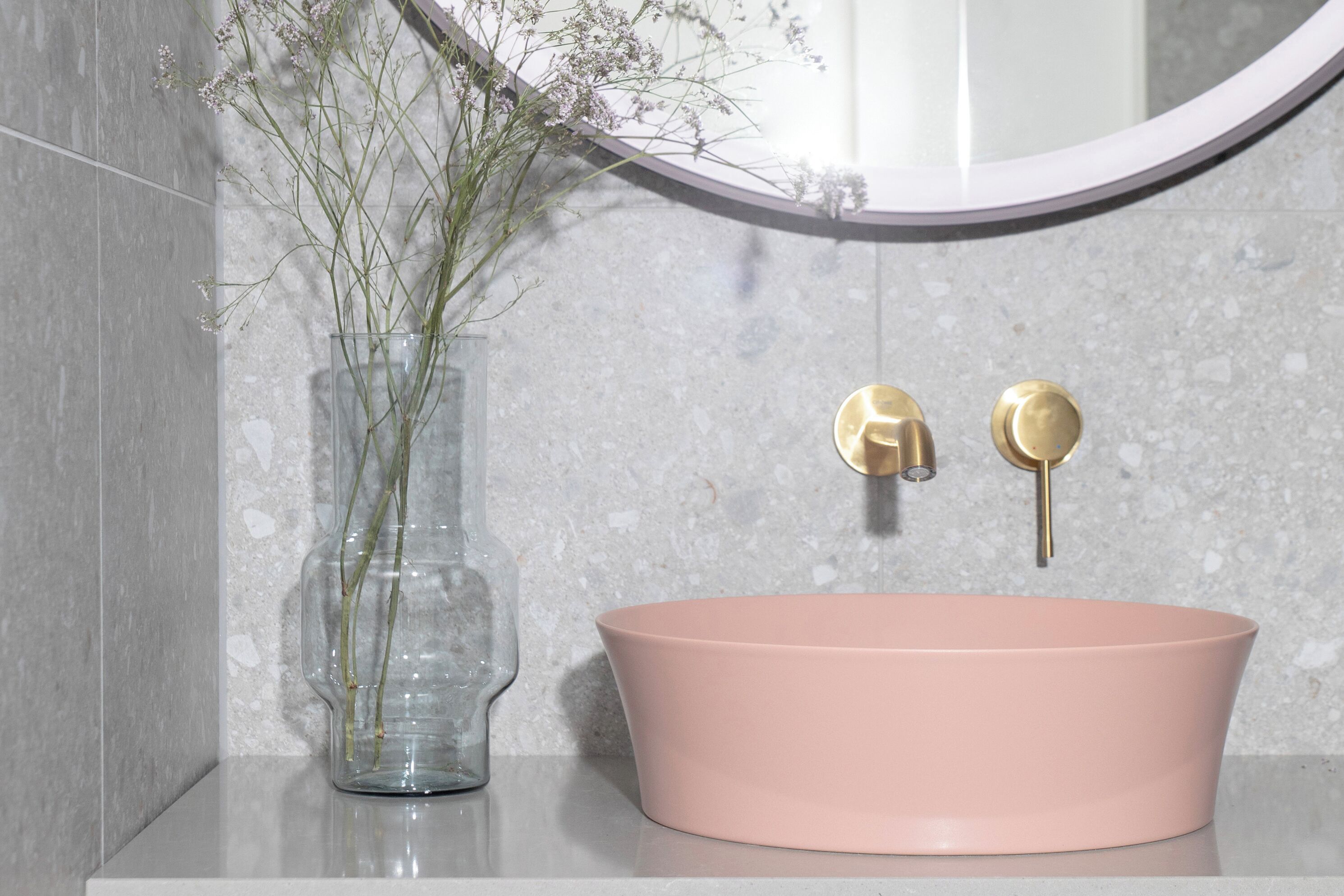 A bathroom with a pink sink, a clear vase with flowers, and a round mirror.