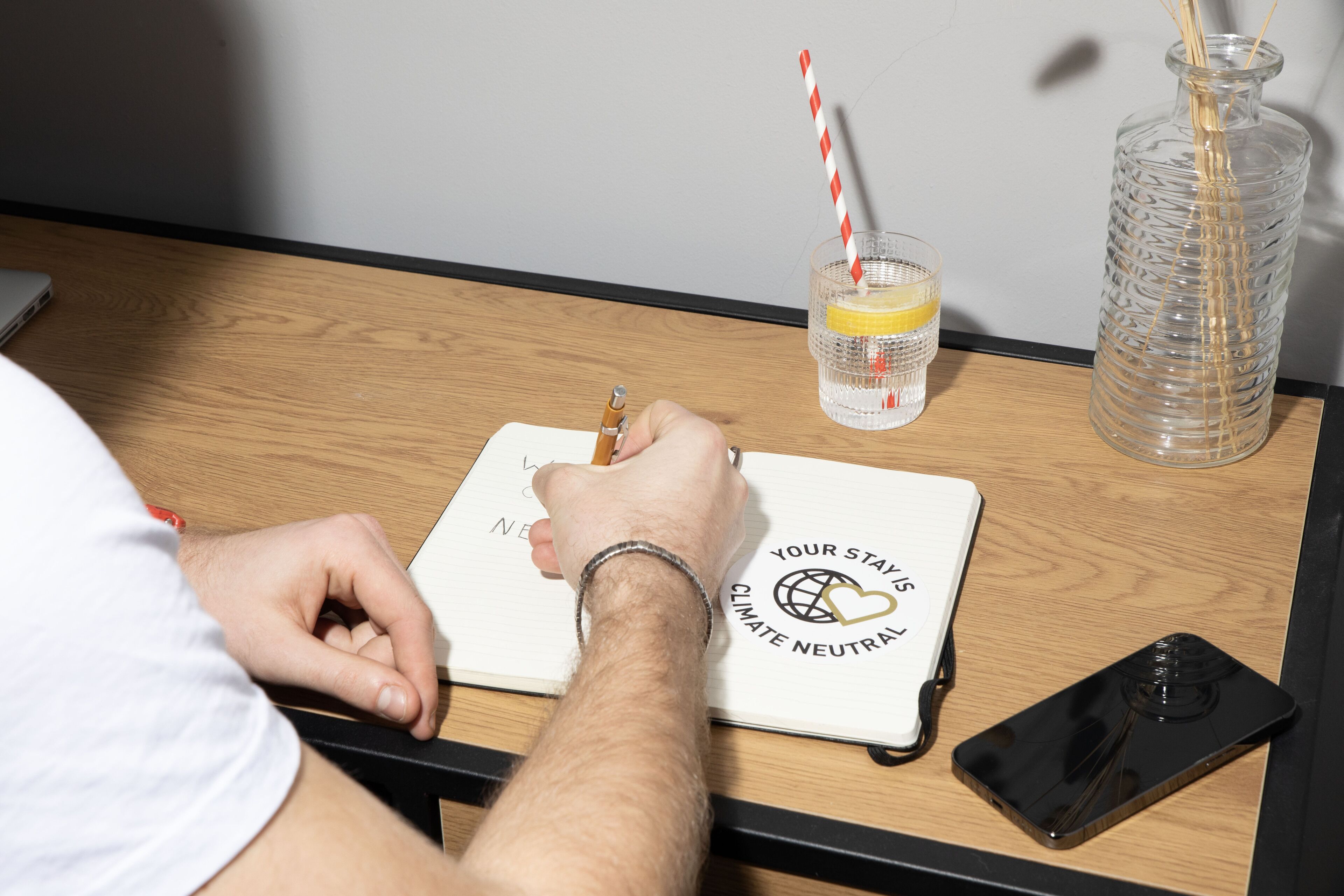 Una persona escribe en un cuaderno con un bolígrafo, un vaso de agua con pajita y un teléfono inteligente sobre una mesa de madera.