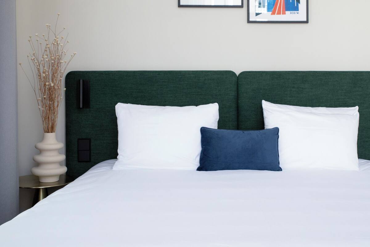 A neatly made bed with white and blue pillows, a green headboard, and a vase with dried plants on a bedside table.