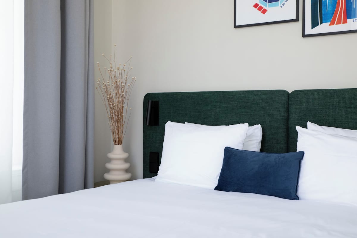 A neatly made bed with white and blue pillows, a green headboard, and a white vase with dried plants on a white nightstand.