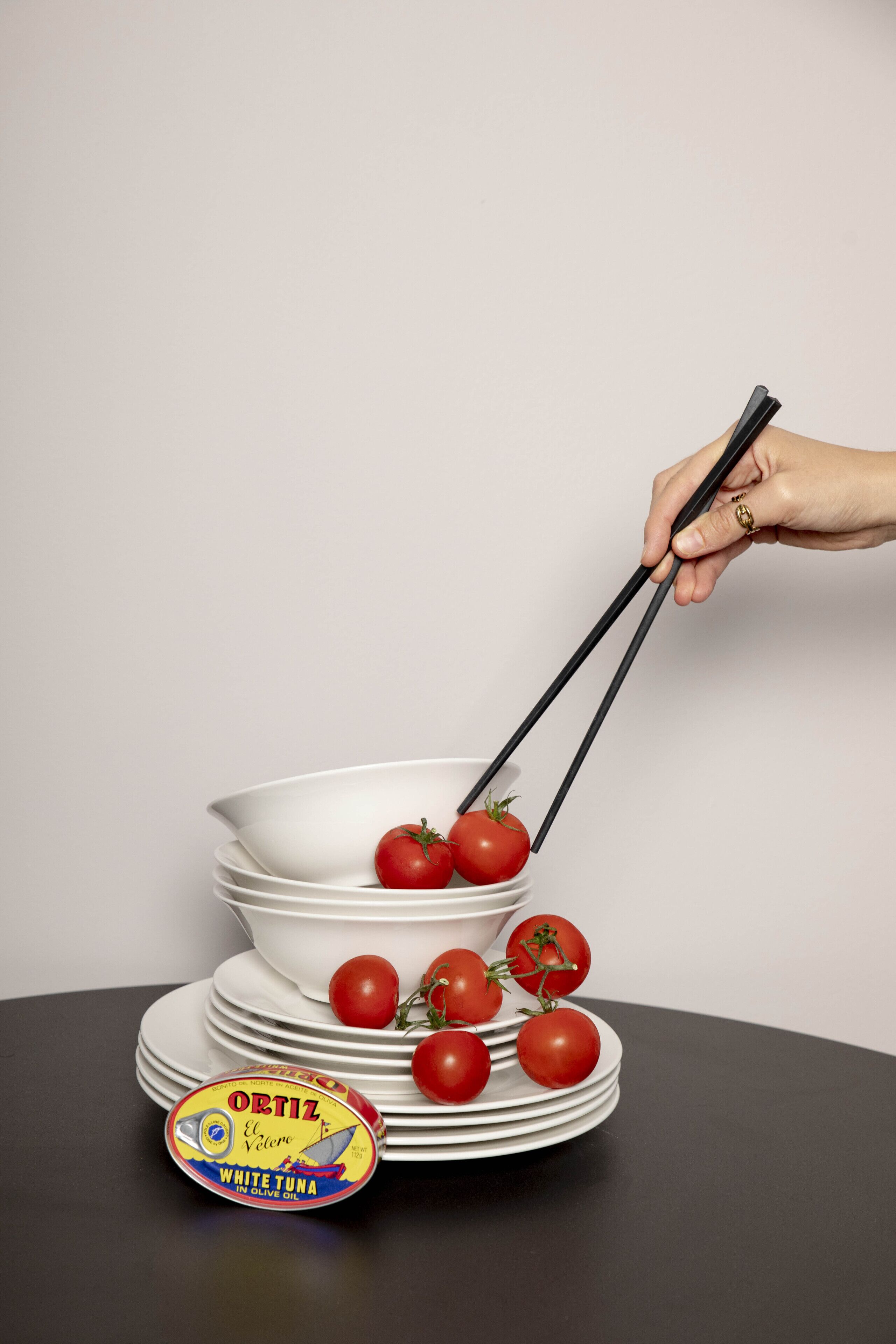 Une main tenant des baguettes au-dessus d’une pile de bols blancs contenant des tomates rouges, le tout posé sur une table noire.