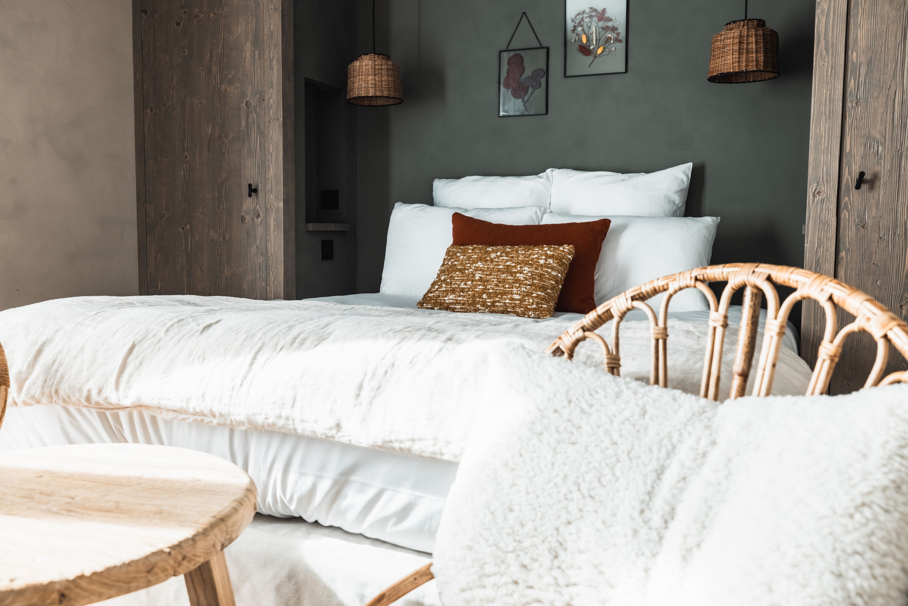 Una camera da letto con un letto grande, una poltrona in vimini, un tavolino rotondo in legno e una parete grigia decorata con opere d'arte.