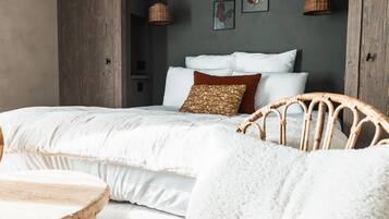 Una camera da letto con un letto grande, una poltrona in vimini, un tavolino rotondo in legno e una parete grigia decorata con opere d'arte.