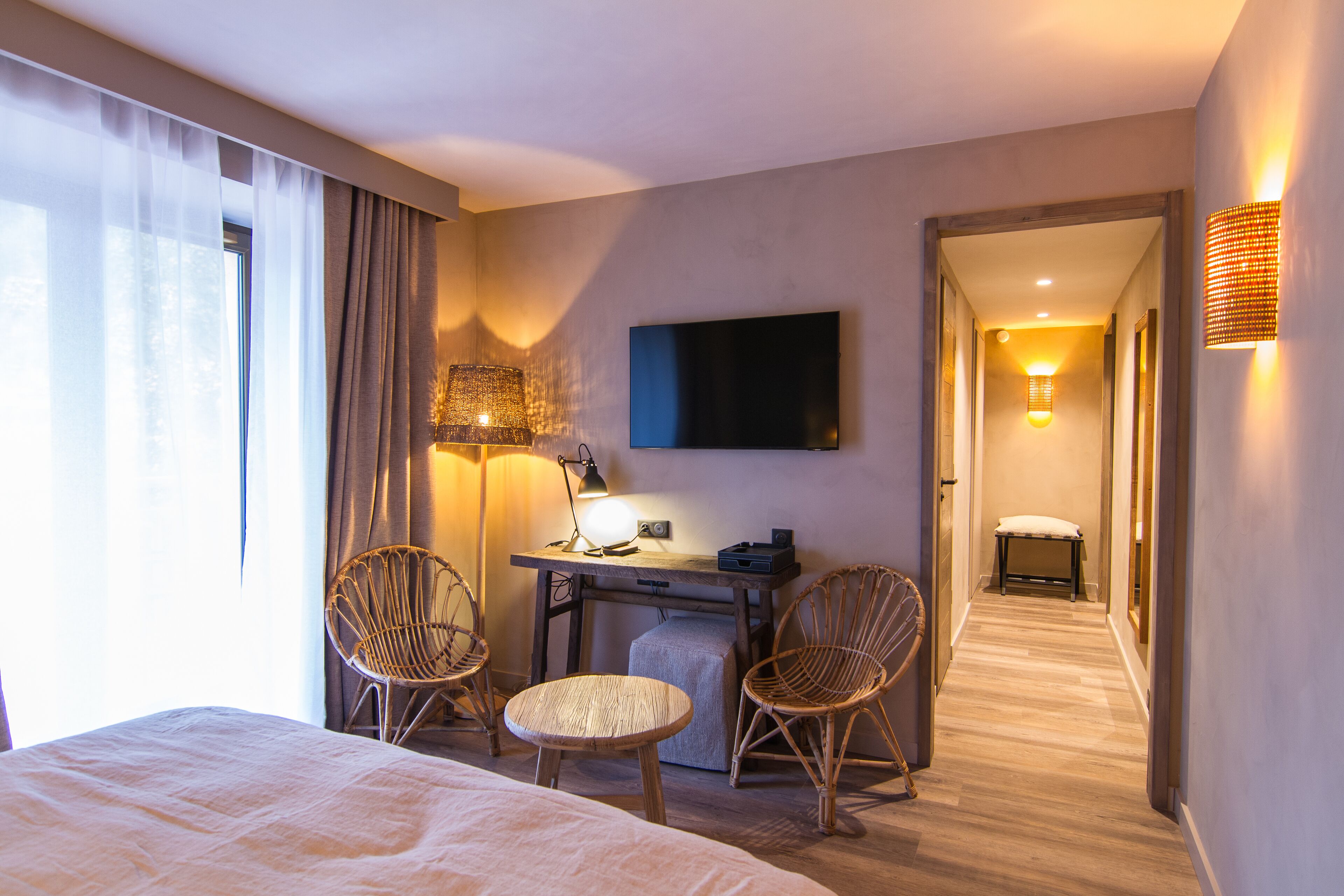 Quarto de hotel com cama, duas cadeiras de vime, escrivaninha com abajur, televisão de tela plana e corredor com um banco.