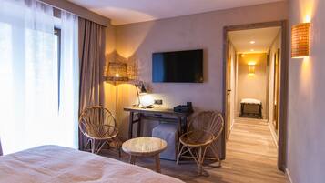 Quarto de hotel com cama, duas cadeiras de vime, escrivaninha com abajur, televisão de tela plana e corredor com um banco.