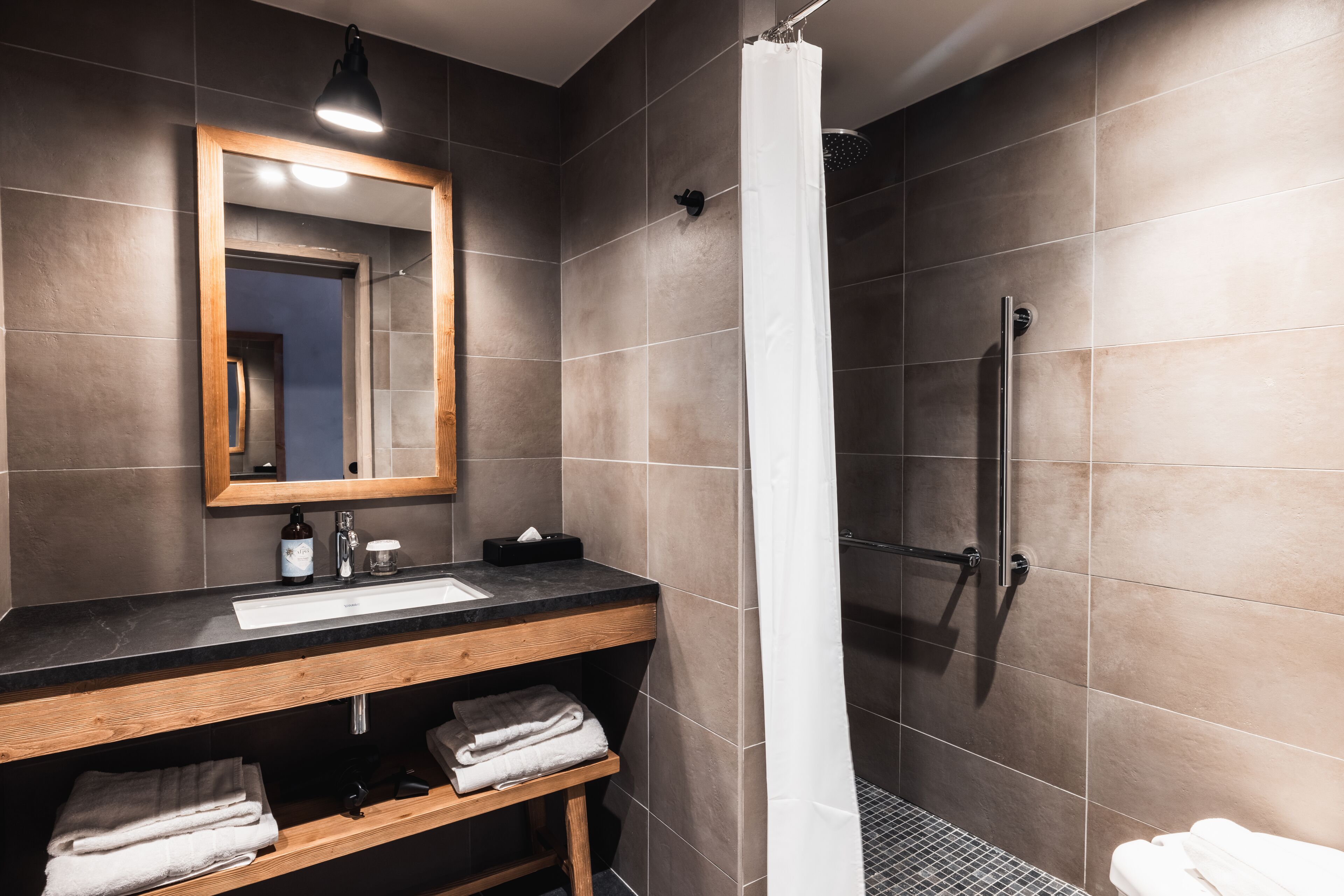 Un bagno moderno con un mobile vanitario in legno, un grande specchio, una doccia con tenda bianca e pareti piastrellate.