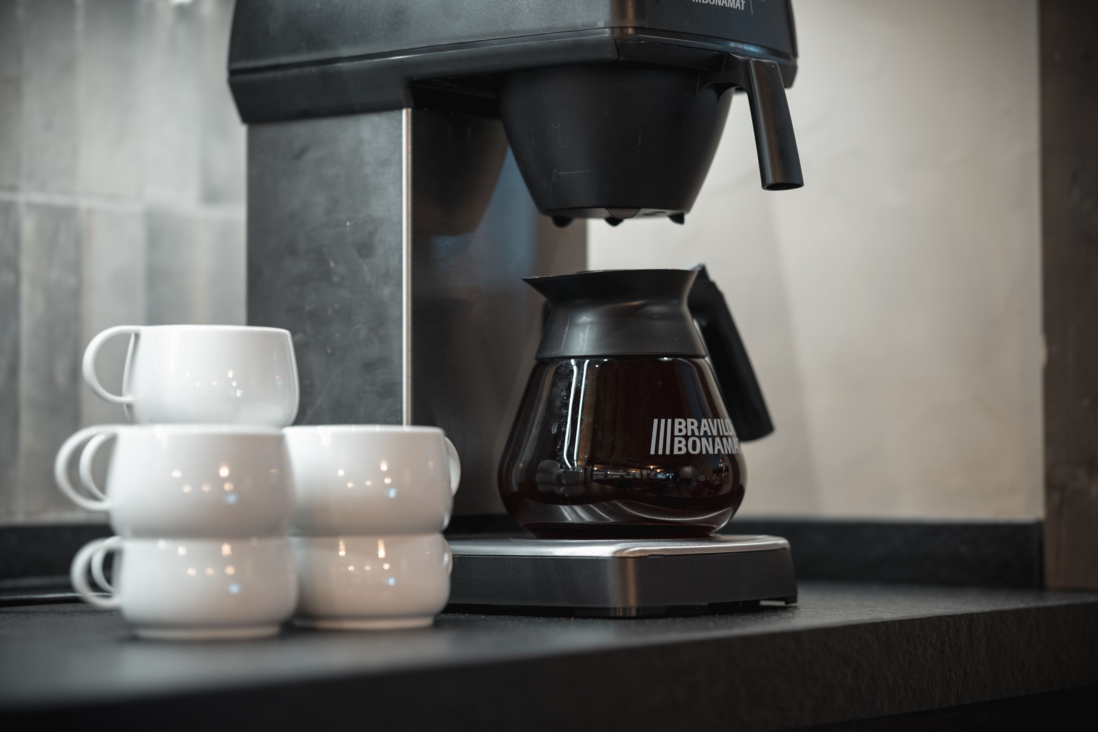 A coffee maker with a branded carafe and four white cups on a countertop.