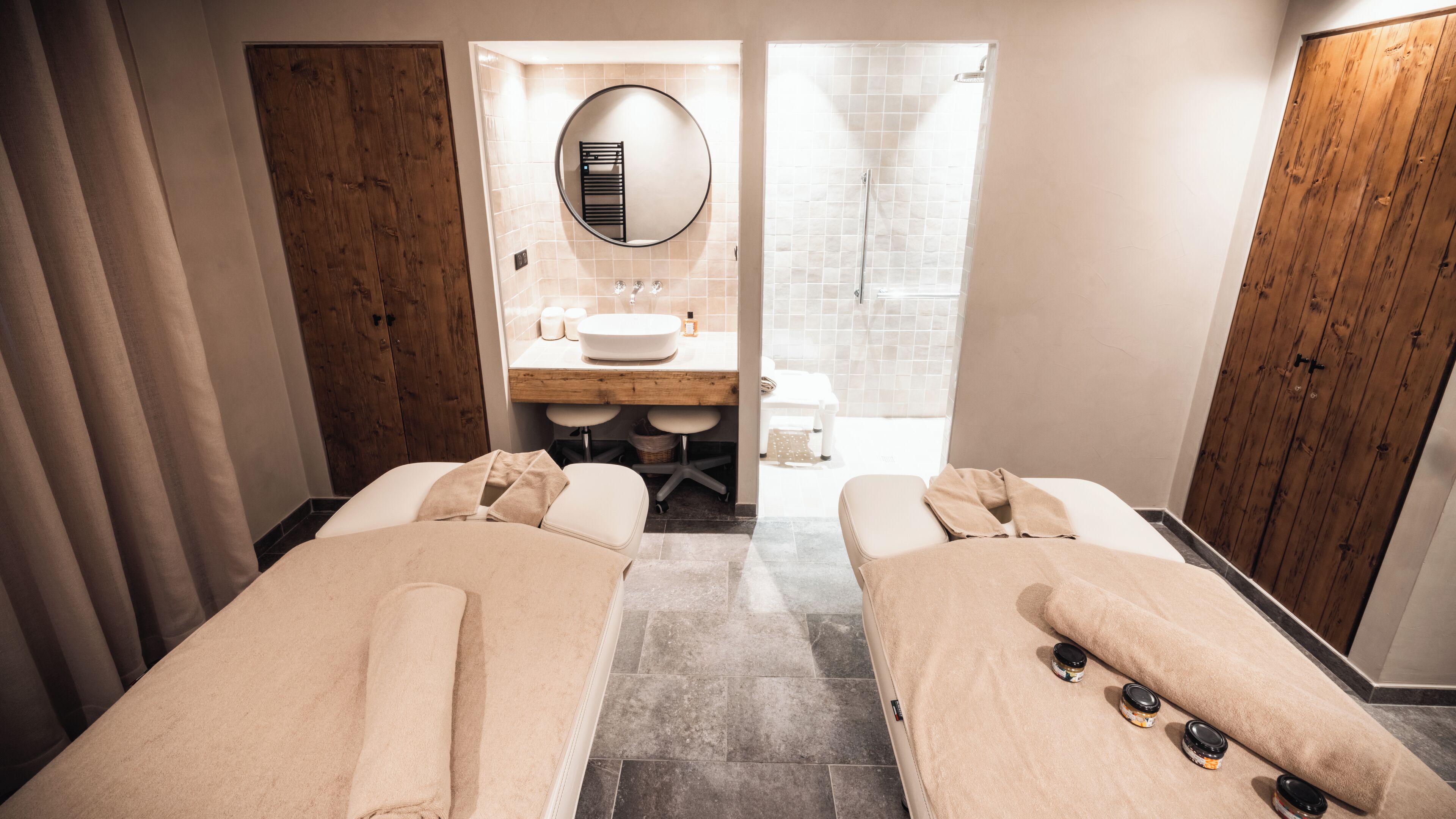 A modern spa room with two massage tables, a wooden vanity, a round mirror, and a shower area.
