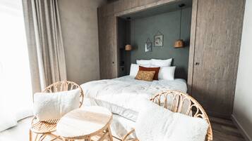 A bedroom with a bed, two wicker chairs, a small round table, and a view of a cityscape through a window.