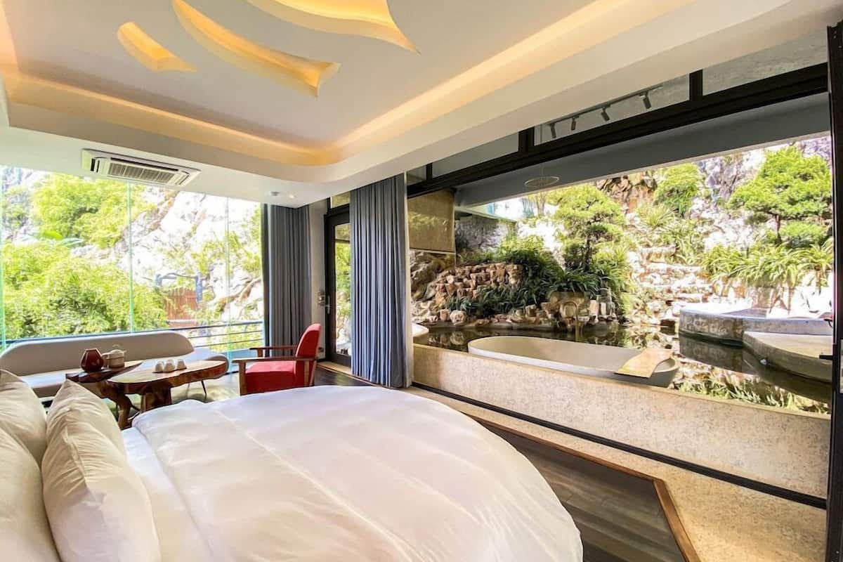 Ein modernes Hotelzimmer mit einem großen Bett, einem kleinen Tisch, einem roten Sessel und Blick auf eine natürliche Landschaft durch ein großes Fenster.