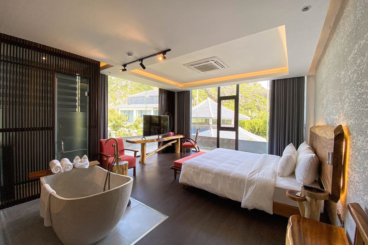 Ein modernes Hotelzimmer mit einer großen Badewanne, einem Bett, einem Schreibtisch mit Fernseher und Blick ins Grüne.