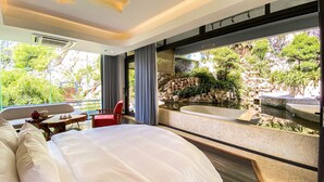 A modern hotel room with a large bed, a small table with chairs, a red chair, and a view of a natural landscape with a waterfall and greenery. - Minawa Kenhga Resort & Spa Ninh Binh (Gia Vien)