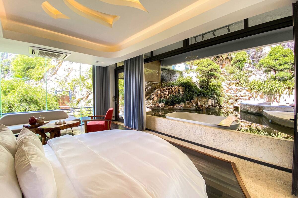 Ein modernes Hotelzimmer mit einem großen Bett, einem kleinen Tisch mit Stühlen, einem roten Sessel und Blick auf eine natürliche Landschaft mit Wasserfall und viel Grün.