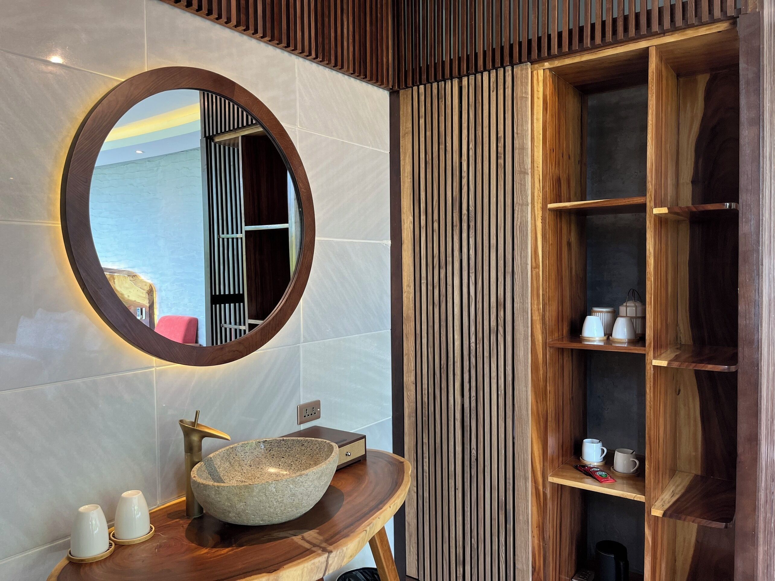 A bathroom with a round mirror, a stone sink, and wooden shelves.