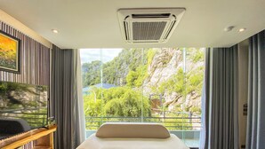 A modern hotel room with a large bed, a wooden coffee table, a TV on a wooden stand, and a balcony with a view of greenery and rocks. - Minawa Kenhga Resort & Spa Ninh Binh (Gia Vien)