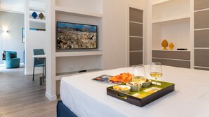 A modern hotel room with a bed, a dining table with a tray of food and drinks, a television on the wall, and a cityscape painting. - La Musa Apartments & Rooms (Palermo)