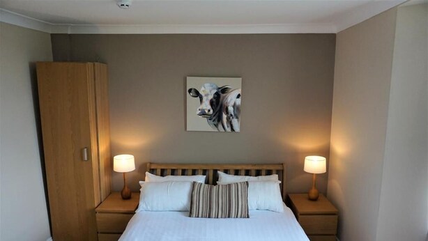 A neatly made bed with white linens, two bedside lamps, a wooden wardrobe, and a cow painting on the wall. - Rooms at The Angel (Cardigan)