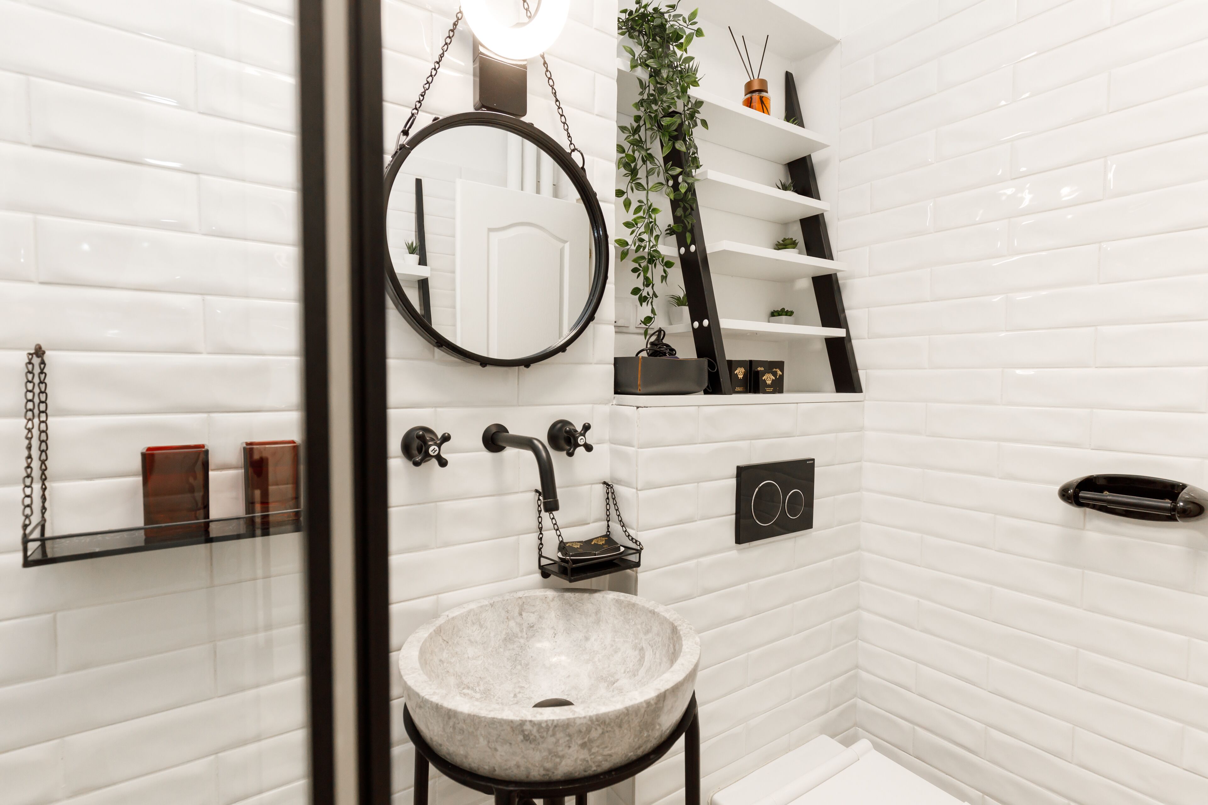 A bathroom with white tiled walls, a round mirror, a marble sink, shelves with toiletries, and a hanging plant.