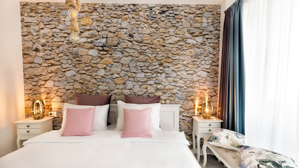 A bedroom with a stone wall, a bed with white bedding and pink pillows, bedside tables with lamps, and a bench with floral cushions. - Metropole Apartments Ateneum Residence (Bucharest)