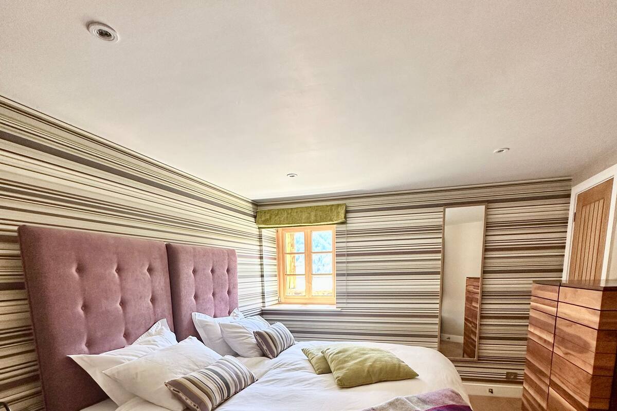 A bedroom with a large bed, striped wall paneling, a window, and a wooden dresser.